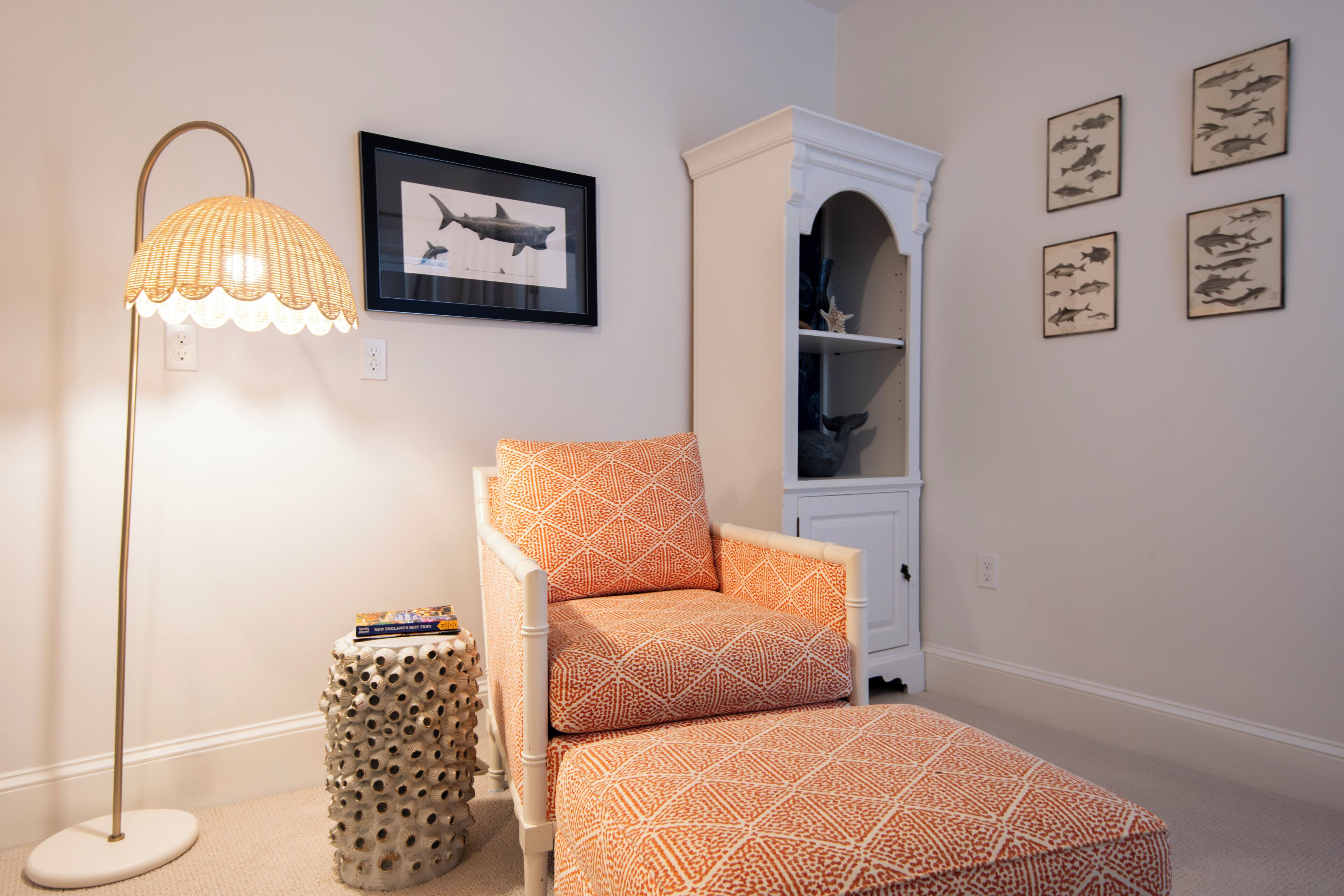 A room with a patterned armchair, a floor lamp, a small side table, a bookshelf, and framed fish illustrations on the wall.