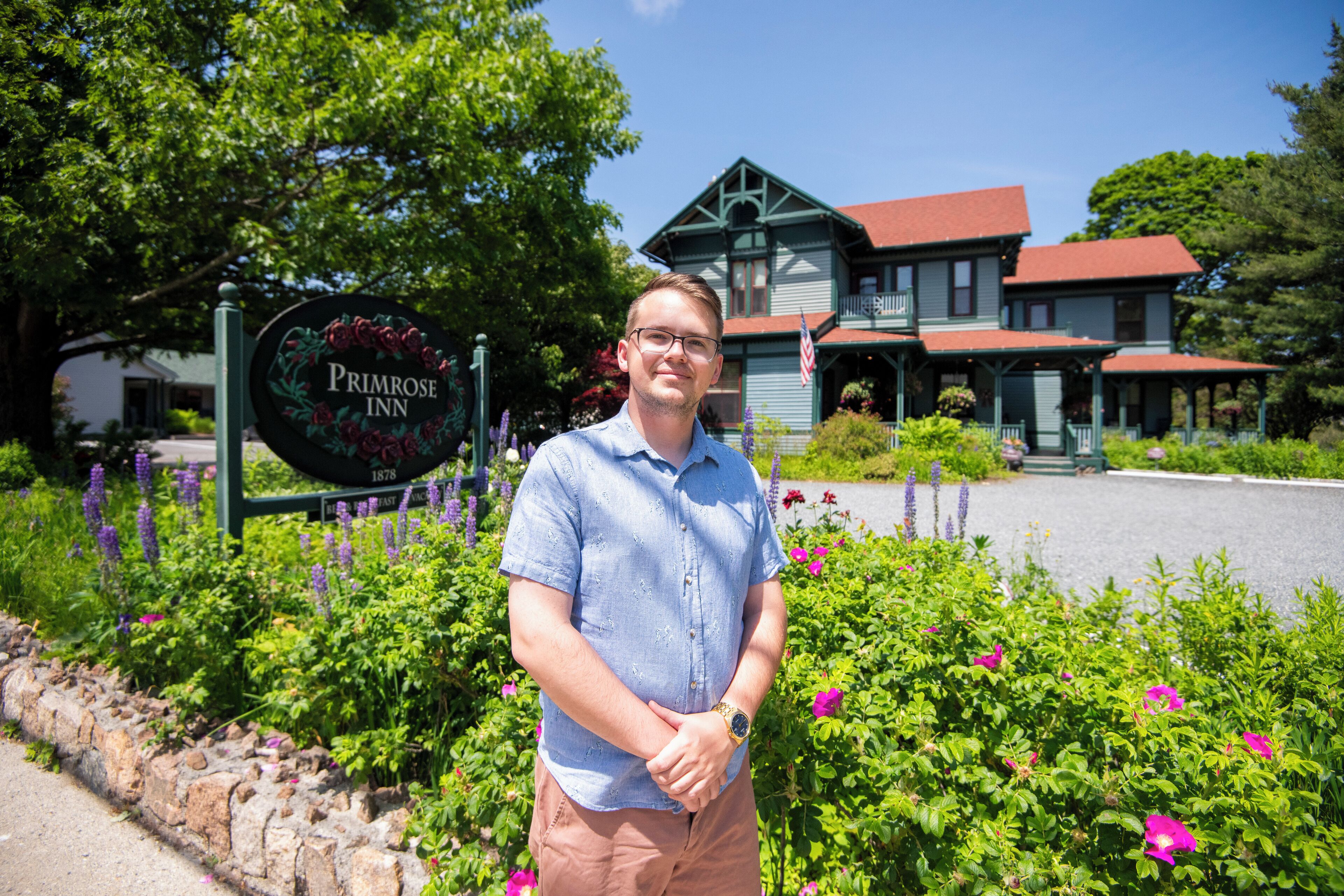 Un homme se tient devant une enseigne indiquant l’emplacement de l’hôtel Primrose Inn, avec une grande maison et une végétation luxuriante en arrière-plan.