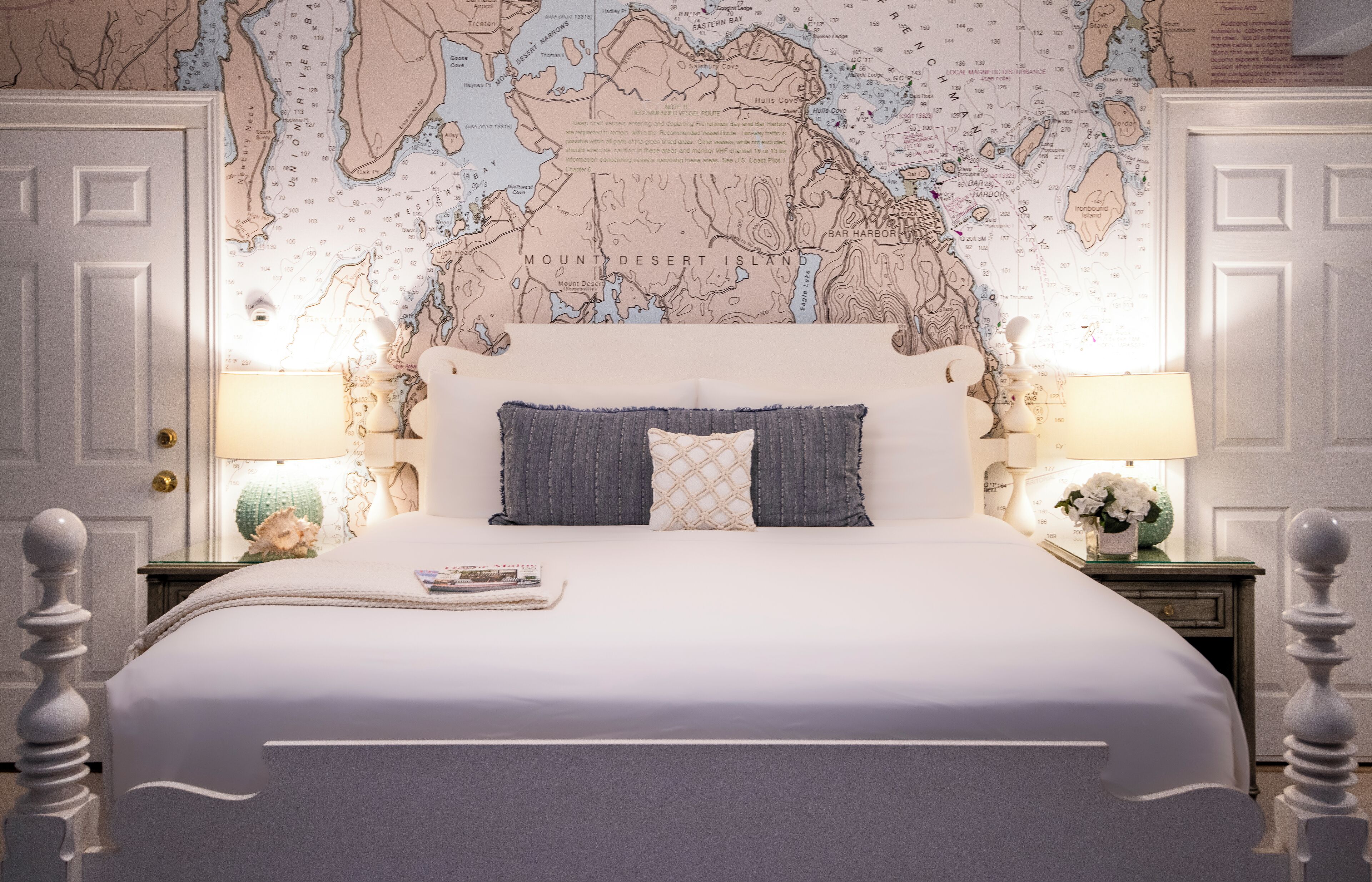 Uma cama bem arrumada com um mapa-múndi como cabeceira, paredes brancas e uma mesinha de cabeceira com abajur e objetos decorativos.