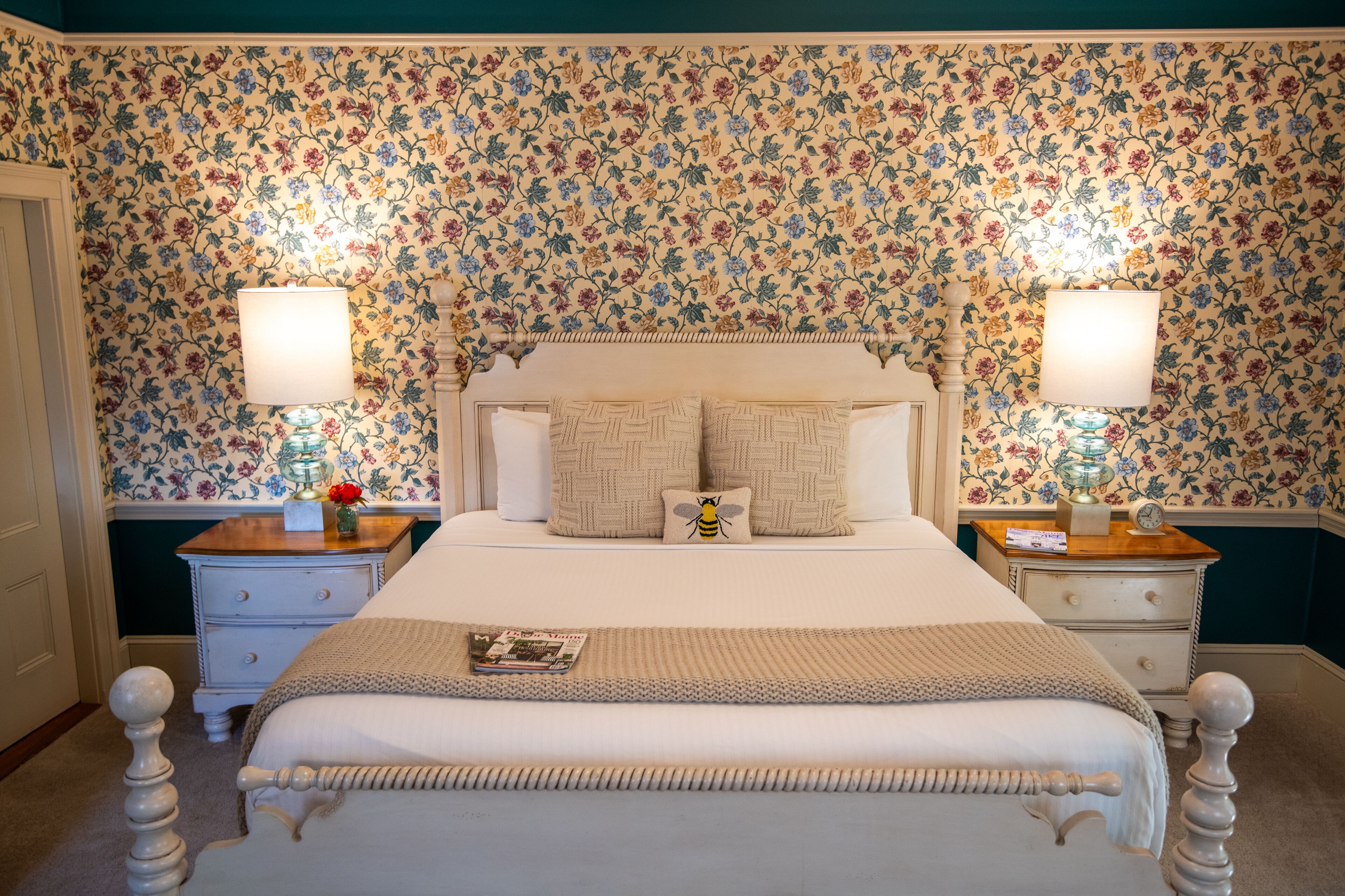 Une chambre à coucher avec un papier peint à motifs floraux, un lit blanc avec une tête de lit, deux tables de chevet avec des lampes et une petite table de nuit avec un vase et une horloge.