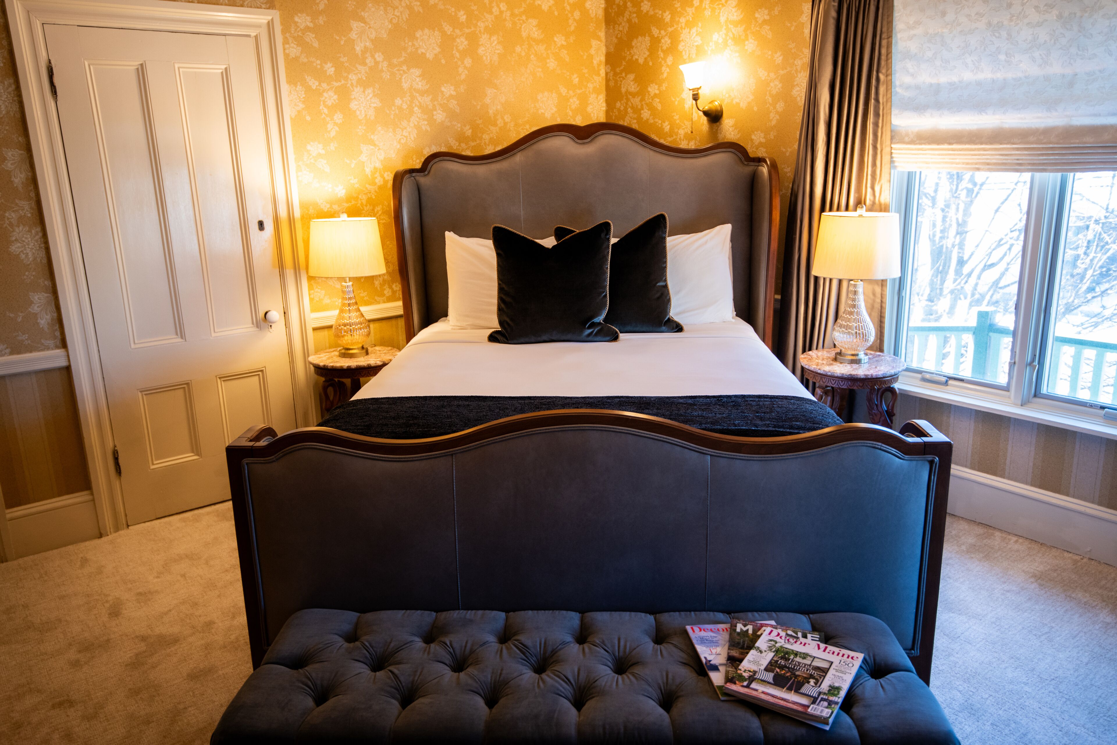 A bedroom with a large bed, two bedside lamps, a window with a view, and a magazine on the footboard.