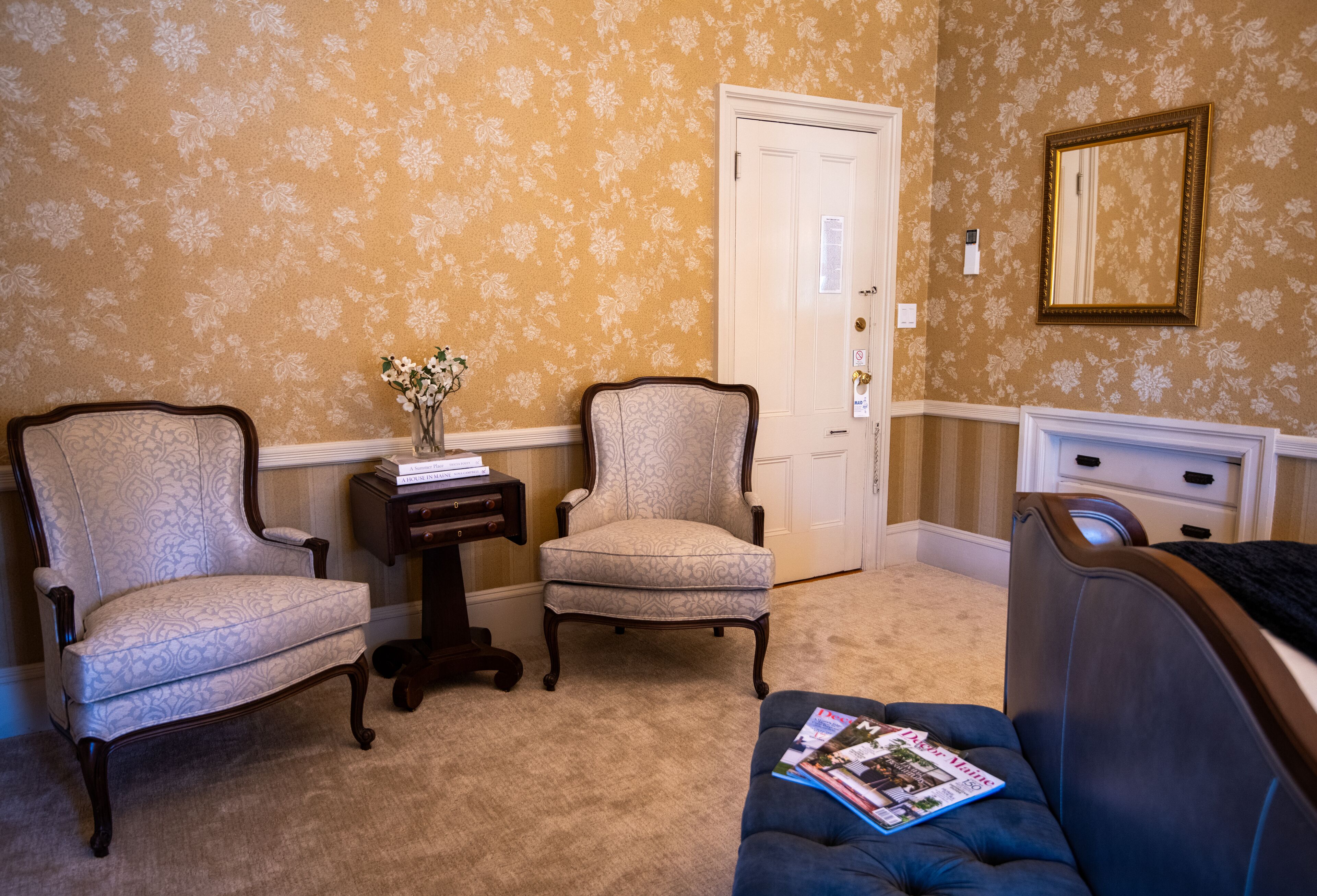 A room with two armchairs, a small table with a vase, a blue sofa with magazines, a door, and a dresser.