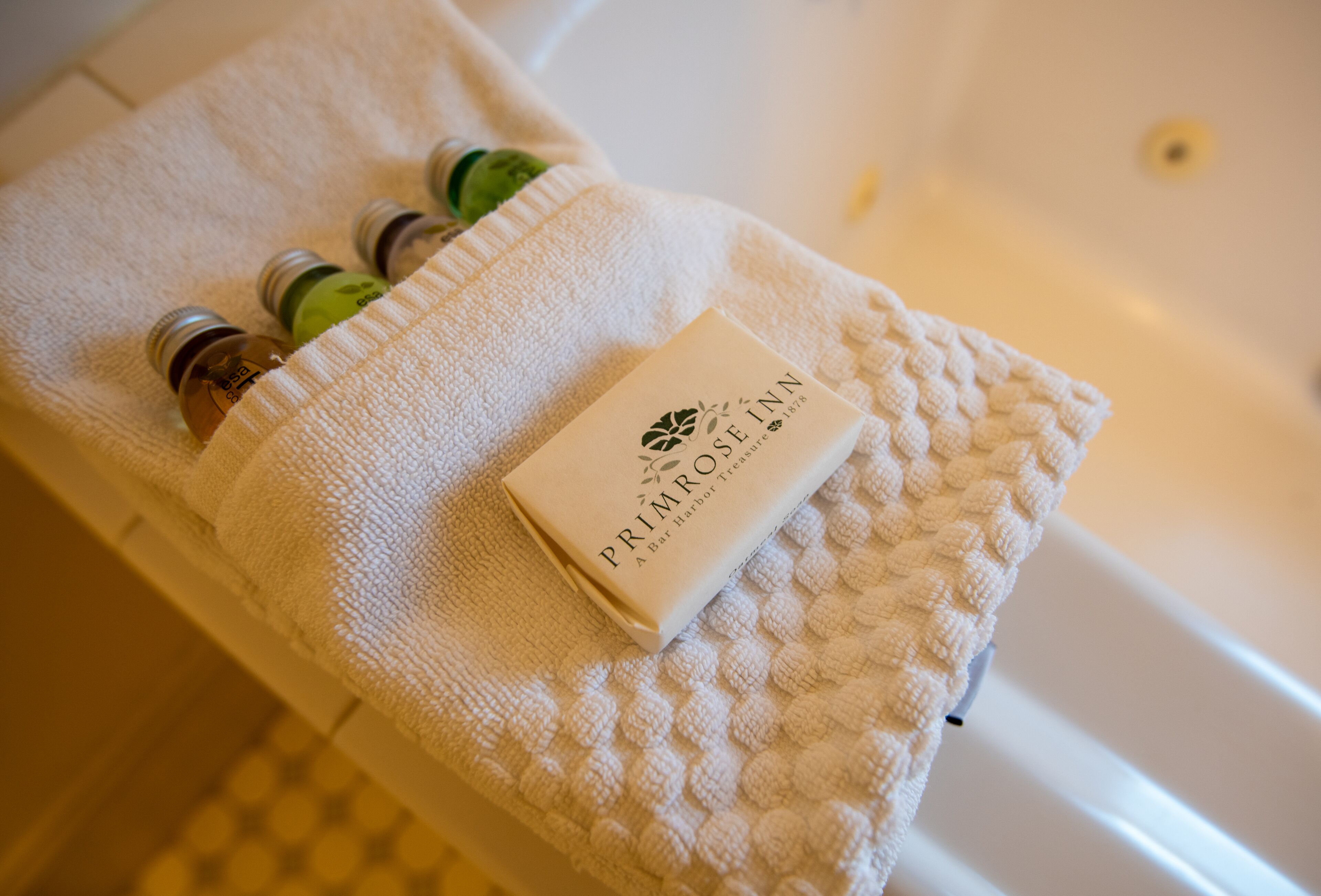 Une serviette blanche avec une étiquette « Primrose Inn » et trois bouteilles sur le bord d’une baignoire.