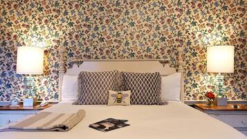 Una cama bien hecha con un edredón a rayas, un cojín con un diseño de abeja y una revista sobre la cama. Hay dos mesitas de noche, con lámparas y un reloj en una de ellas. La habitación tiene papel pintado con motivos florales y un cabecero con un diseño decorativo.