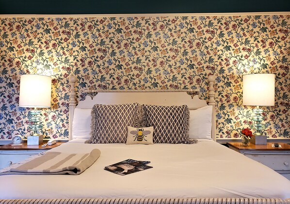 A neatly made bed with a striped blanket, a pillow with a bee design, and a magazine on the bed. There are two bedside tables with lamps and a clock on one of them. The room has floral wallpaper and a headboard with a decorative pattern.