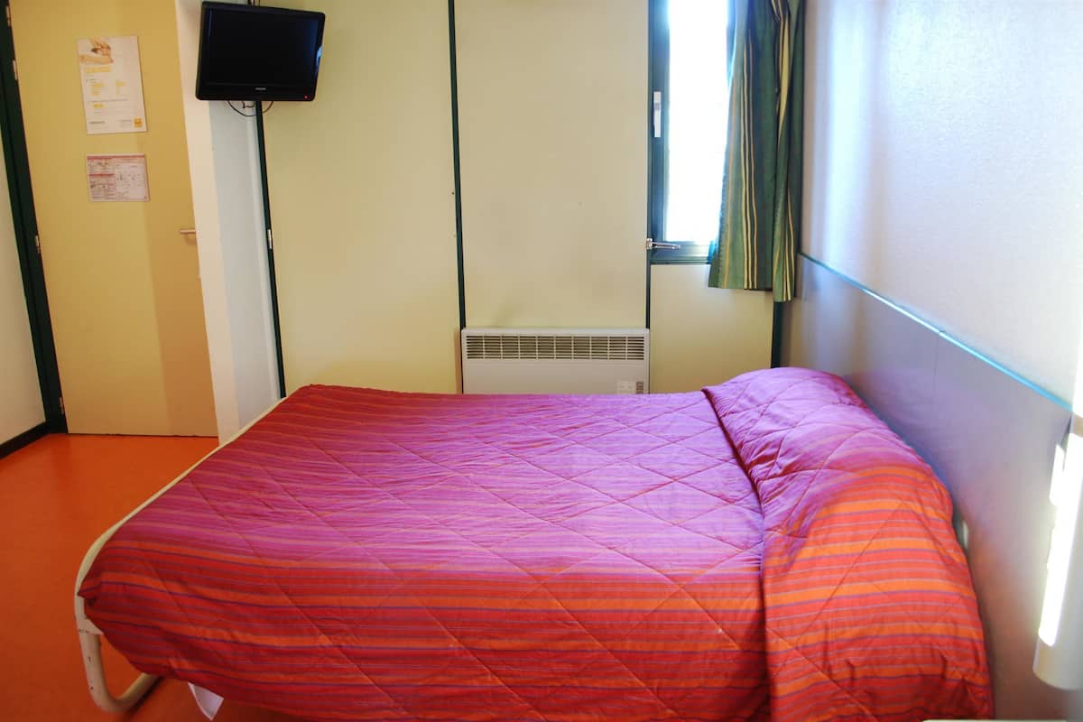 Un lit simple avec une couverture aux couleurs vives, un téléviseur à écran plat fixé au mur et une fenêtre avec des rideaux.