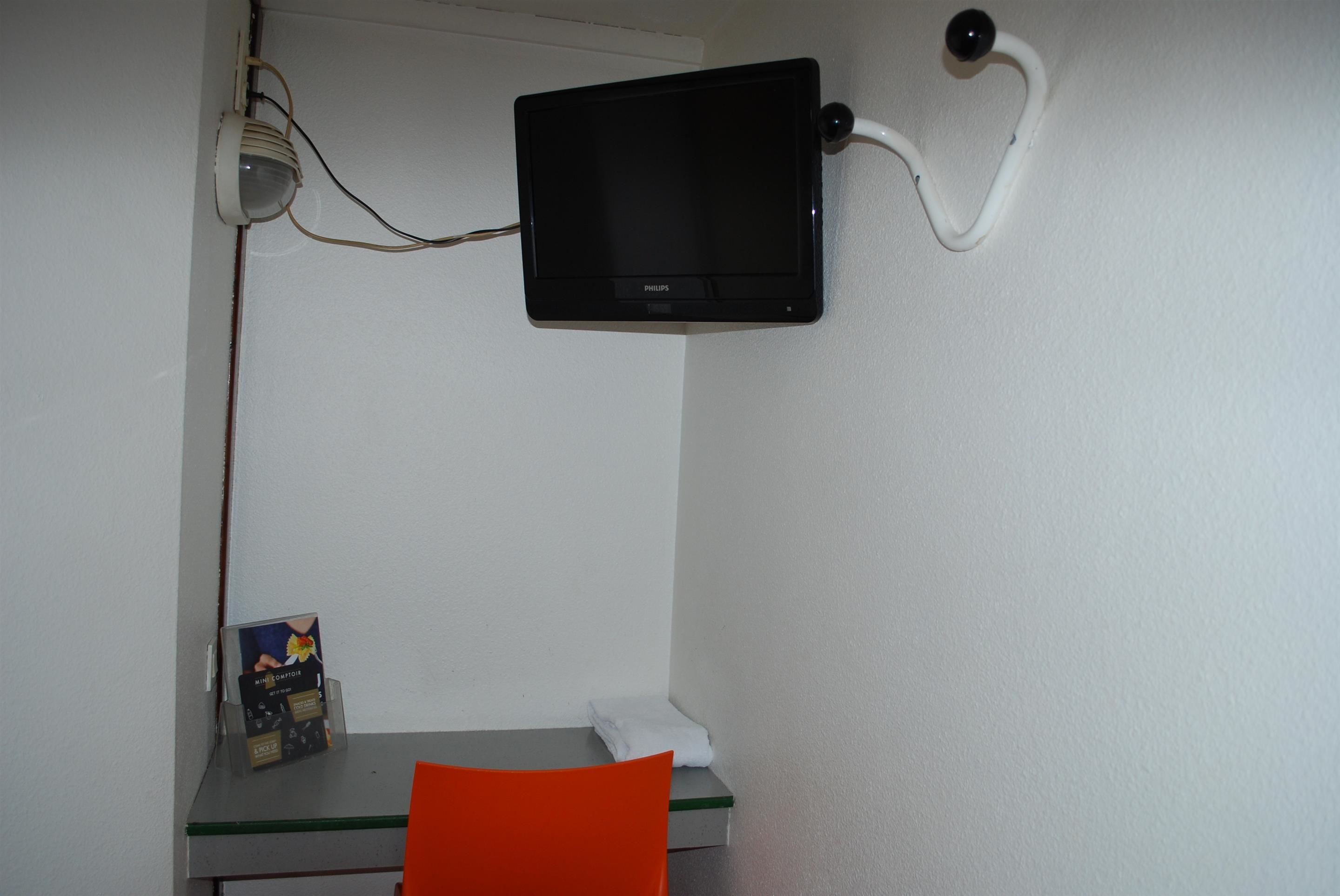 Une petite pièce comprenant un bureau, une chaise, un écran fixé au mur et une petite étagère avec un livre et une boîte de mouchoirs.