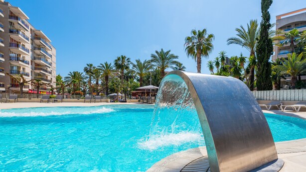Une piscine agrémentée d’un élément décoratif aquatique, entourée de palmiers et bordée d’un bâtiment doté de balcons.