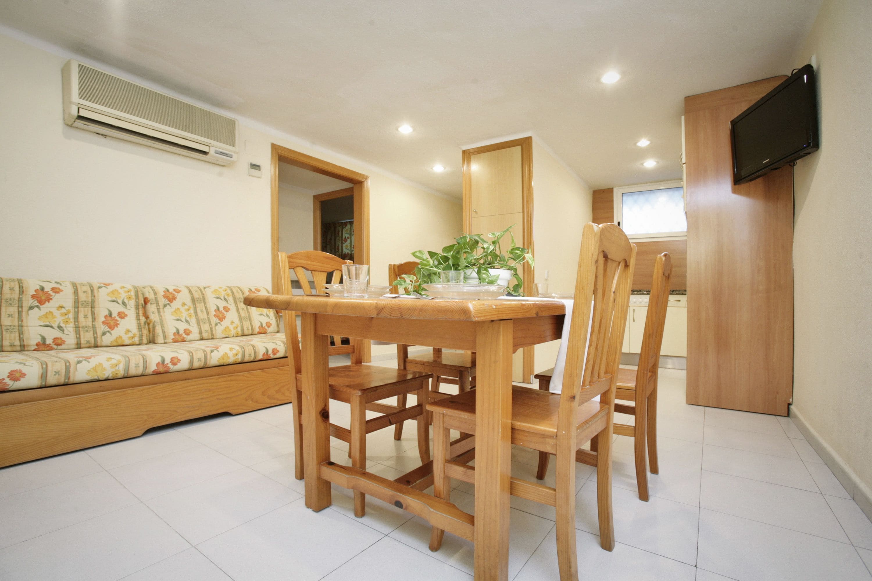 Un coin repas avec une table et des chaises en bois, un canapé recouvert d’un tissu à motifs floraux et un téléviseur fixé au mur.