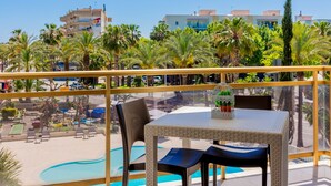 Un balcon avec une table et des chaises donnant sur une piscine, entouré de palmiers et d’immeubles.