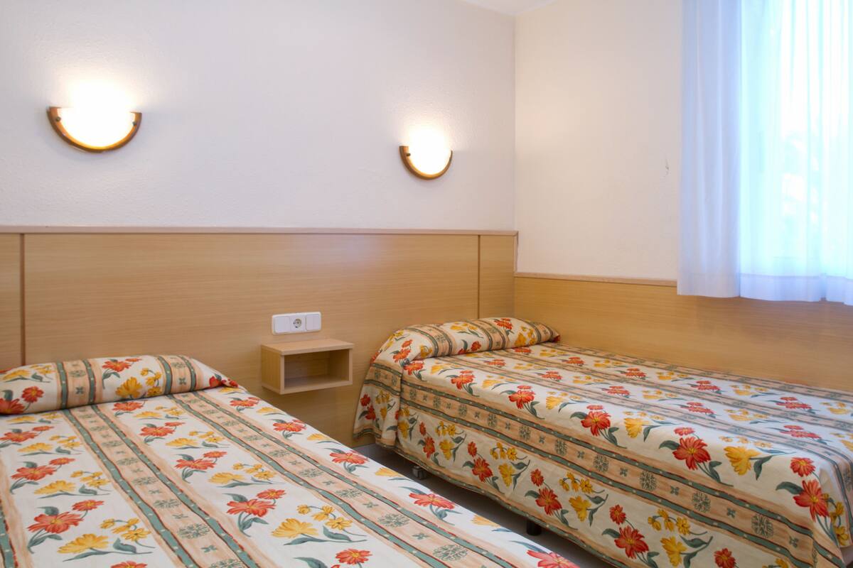 Habitación de hotel con dos camas individuales, cabecera de madera, luces fijadas a la pared y una ventana con cortinas.