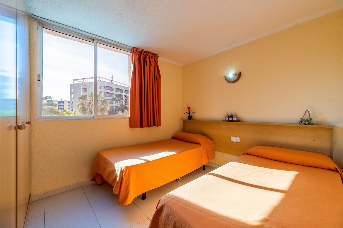 A hotel room with two single beds, a window with curtains, a wall-mounted light, and a small shelf with items.