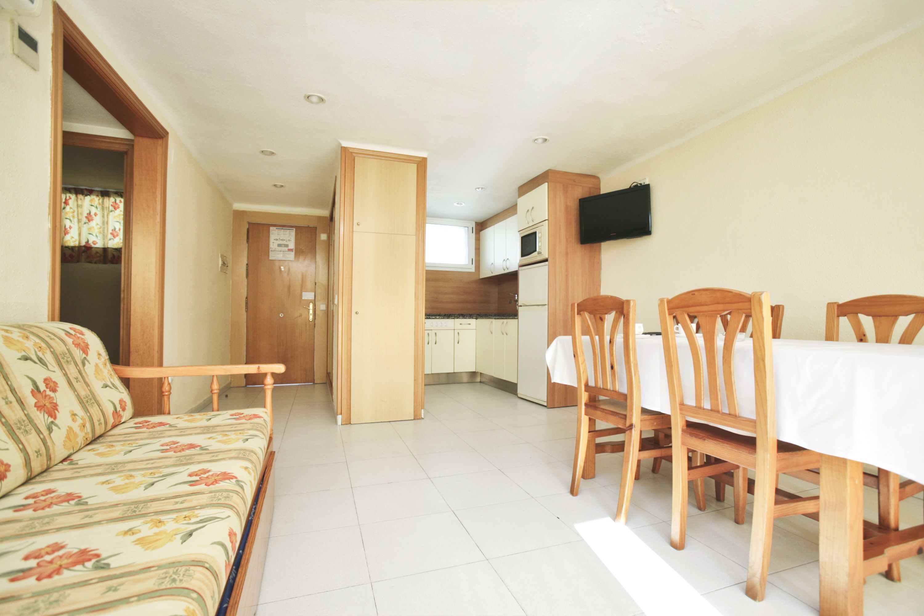 Un espacio habitable compacto con sofá, mesa de comedor, sillas, zona de cocina y televisor montado en la pared.