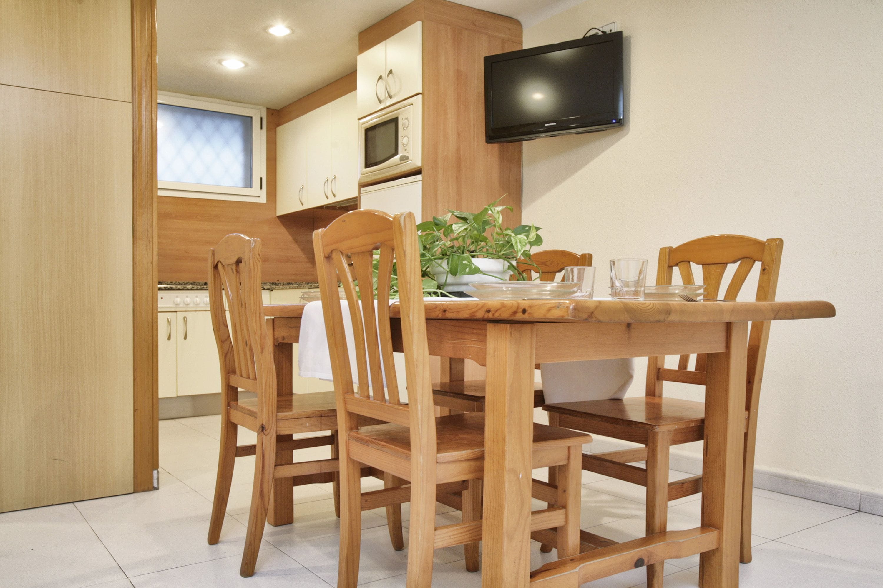 A wooden dining table with four chairs, a television mounted on the wall, and a kitchen area with cabinets and a microwave.