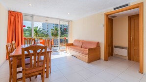 Un vaste espace de vie comprenant une table et des chaises en bois, un canapé et une porte-fenêtre coulissante donnant sur un balcon avec vue sur les palmiers et les immeubles.