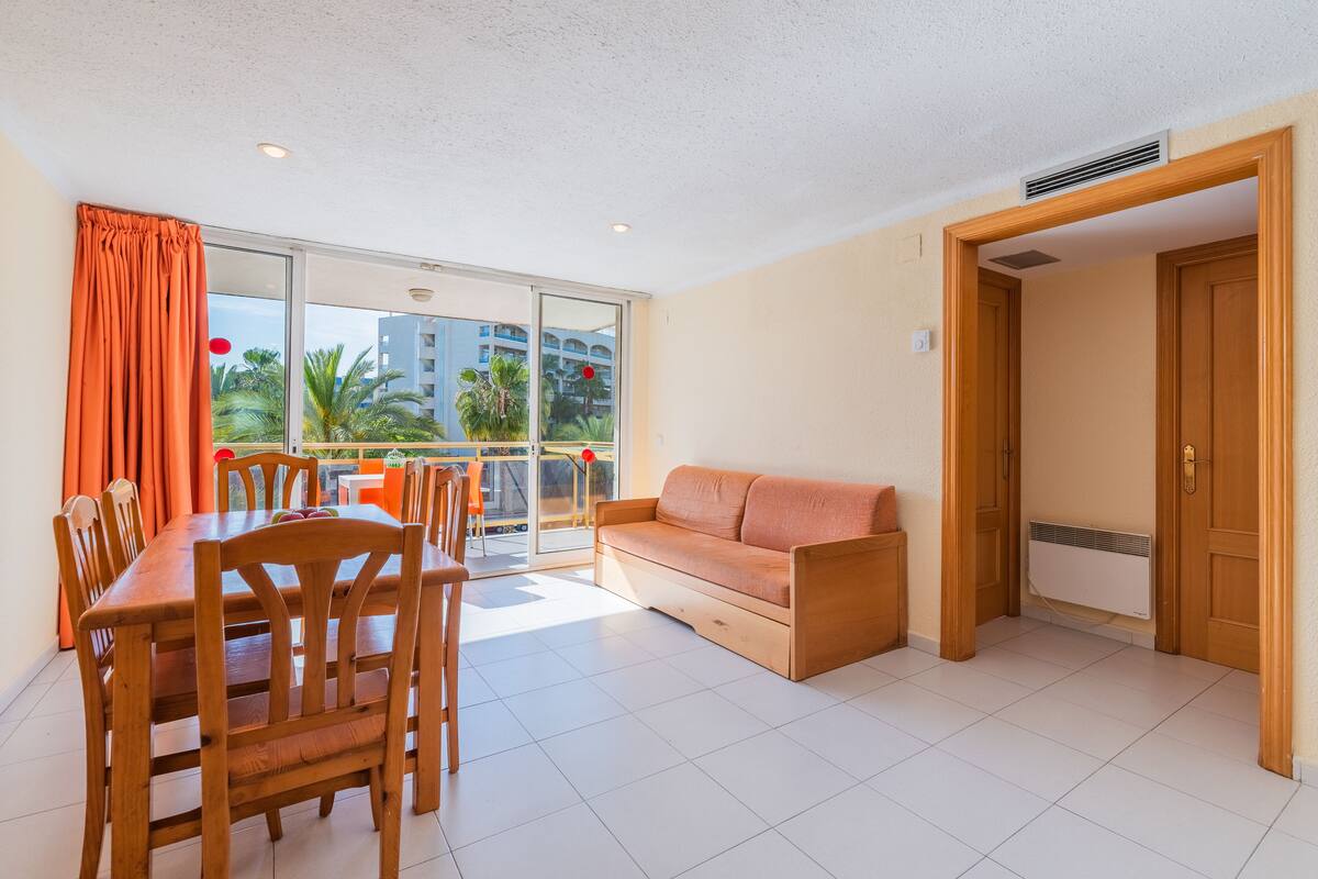 Amplia sala de estar con una mesa y sillas de madera, un sofá y un ventanal corredero que da a un balcón con vistas a palmeras y edificios.