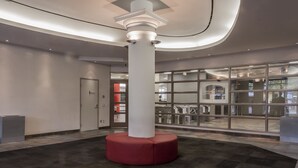 Um hall amplo com uma coluna central, uma área de recepção e vista para a livraria da faculdade através de portas de vidro.
