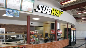 Um restaurante Subway com painel de cardápio, vitrine de alimentos e refrigerador de Coca-Cola.