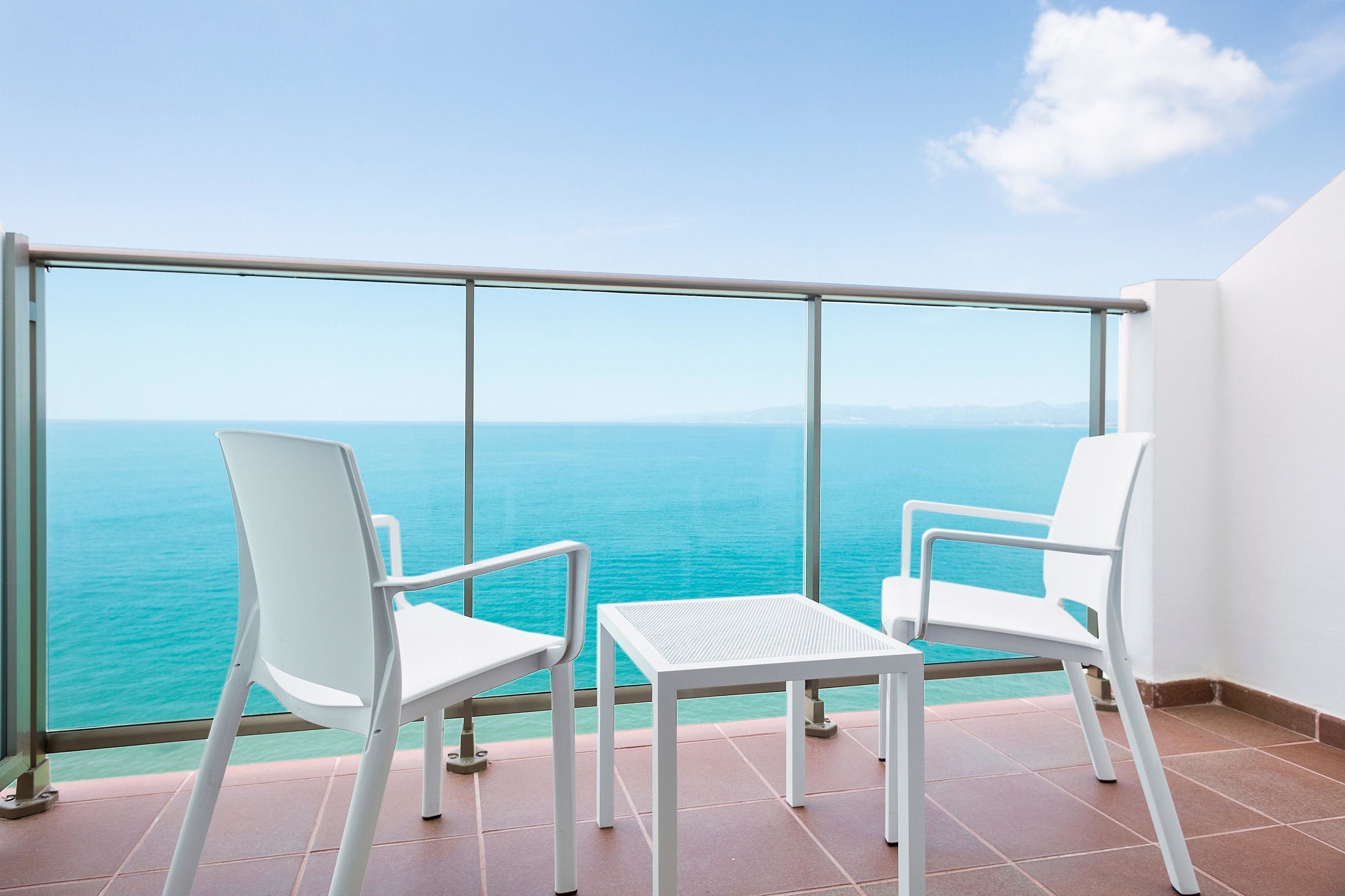 Un balcon avec deux chaises blanches et une petite table, d’où l’on peut admirer une mer turquoise et des montagnes lointaines.