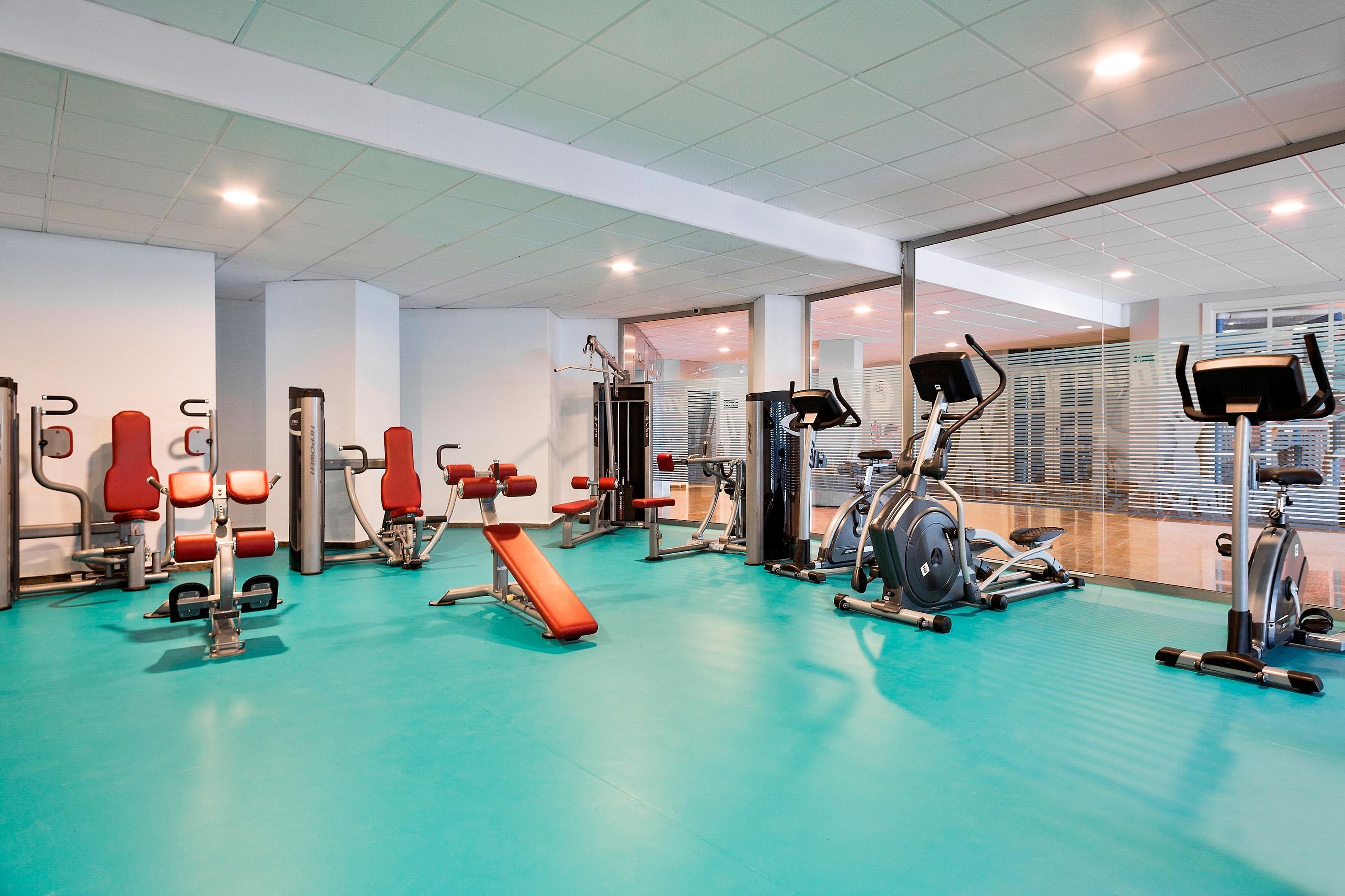 Un gimnasio con diverso equipo de ejercicio, incluyendo cintas de correr y máquinas de pesas, sobre un piso turquesa.