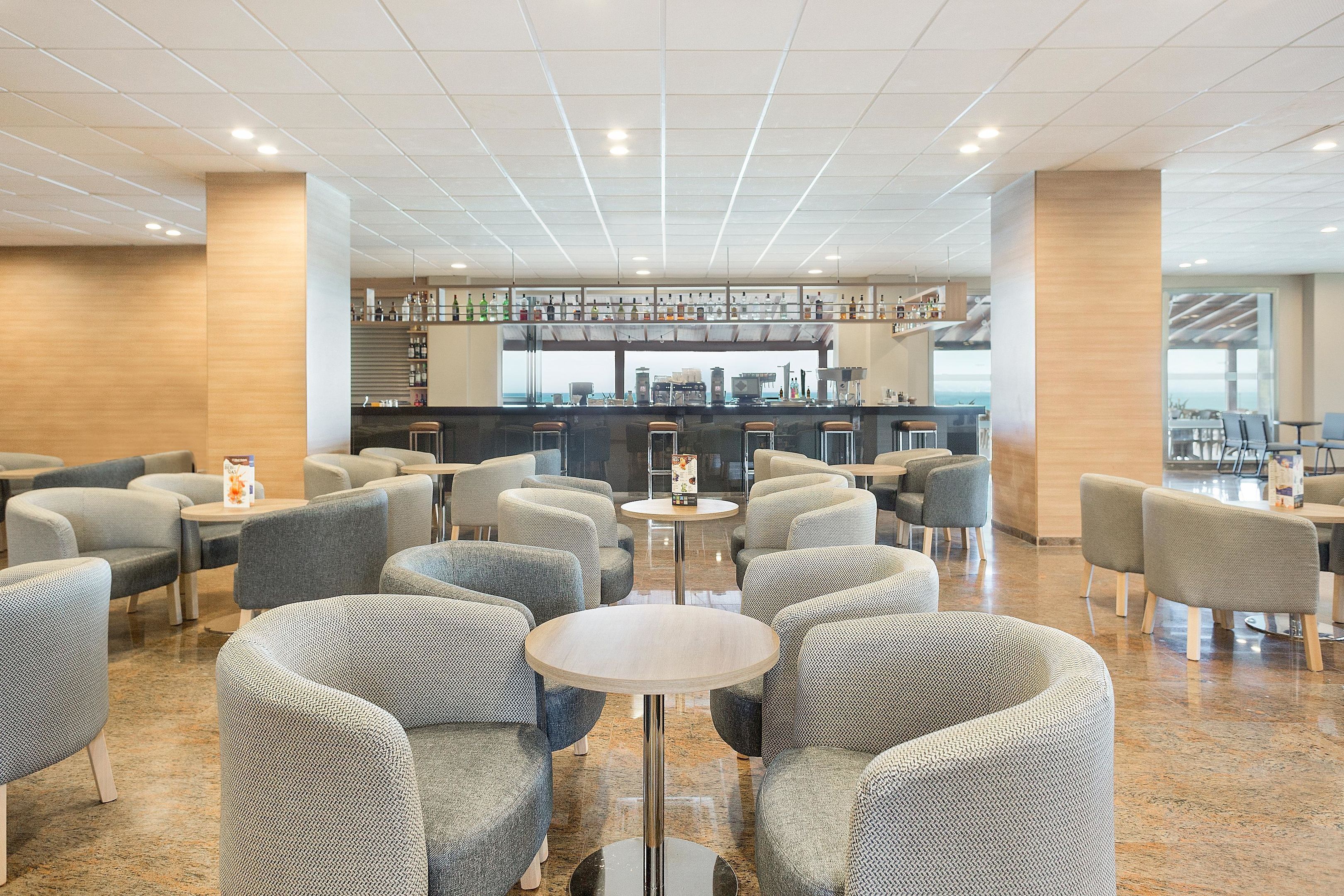 En modern hotellobby med en bardel, sittgrupper och utsikt över havet.