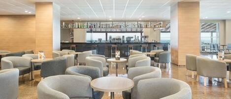 En modern hotellobby med en bardel, sittgrupper och utsikt över havet.