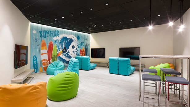 Un espacio interior moderno con un mural de temática surf, coloridos pufs y taburetes de bar.