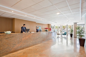 A modern reception area with a marble counter, a receptionist, and a seating area with chairs and a table. - Hotel Best Negresco (Salou)