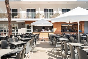 An outdoor café with tables and chairs, a bar area, and a view of the building's upper floor.