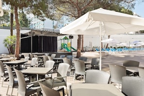 An outdoor seating area with tables and chairs, a stage with a children's play area, and umbrellas providing shade. - Hotel Best Oasis Park (Salou)