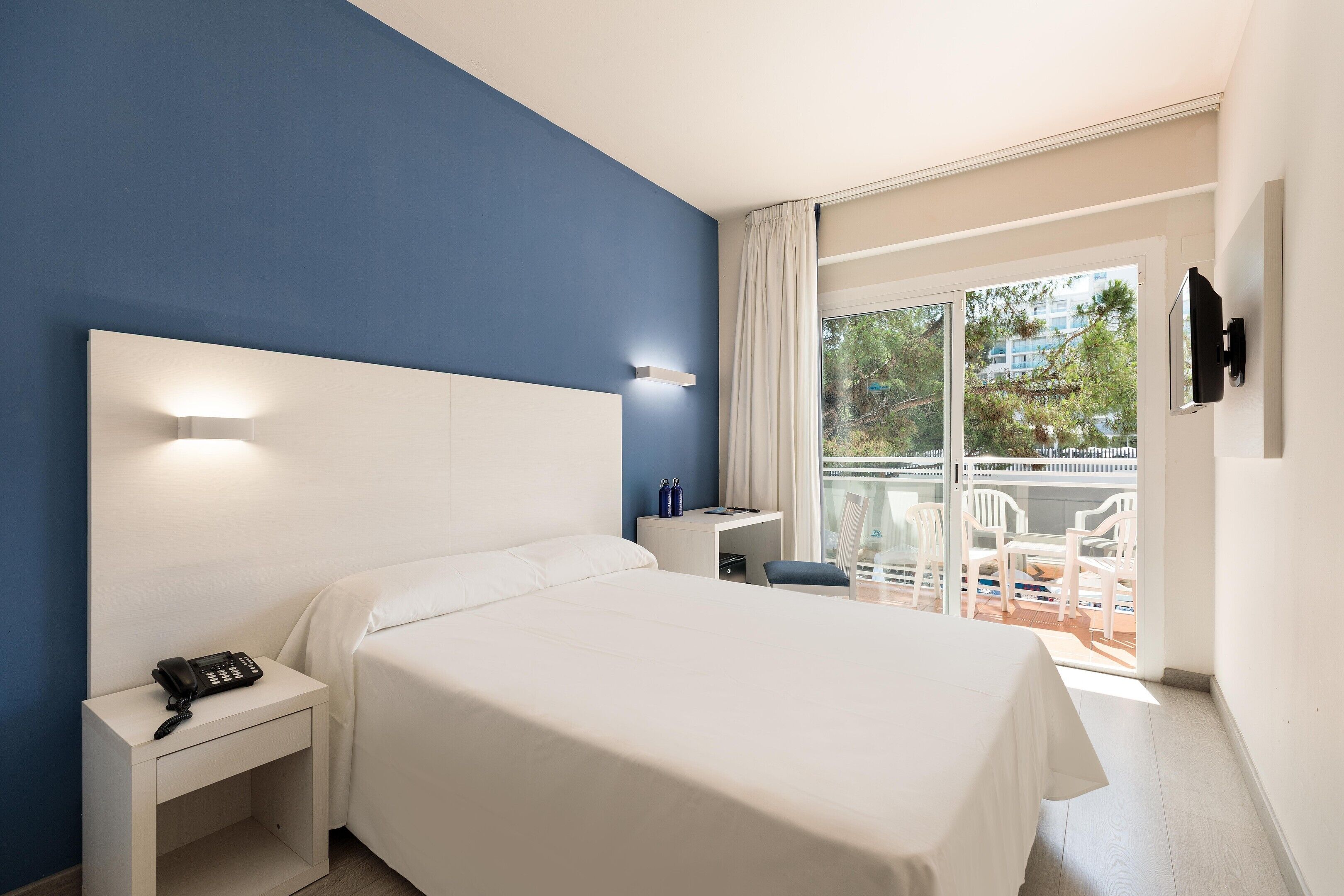Habitación de hotel con cama, escritorio con teléfono, televisor montado en la pared y balcón con sillas y una mesa.