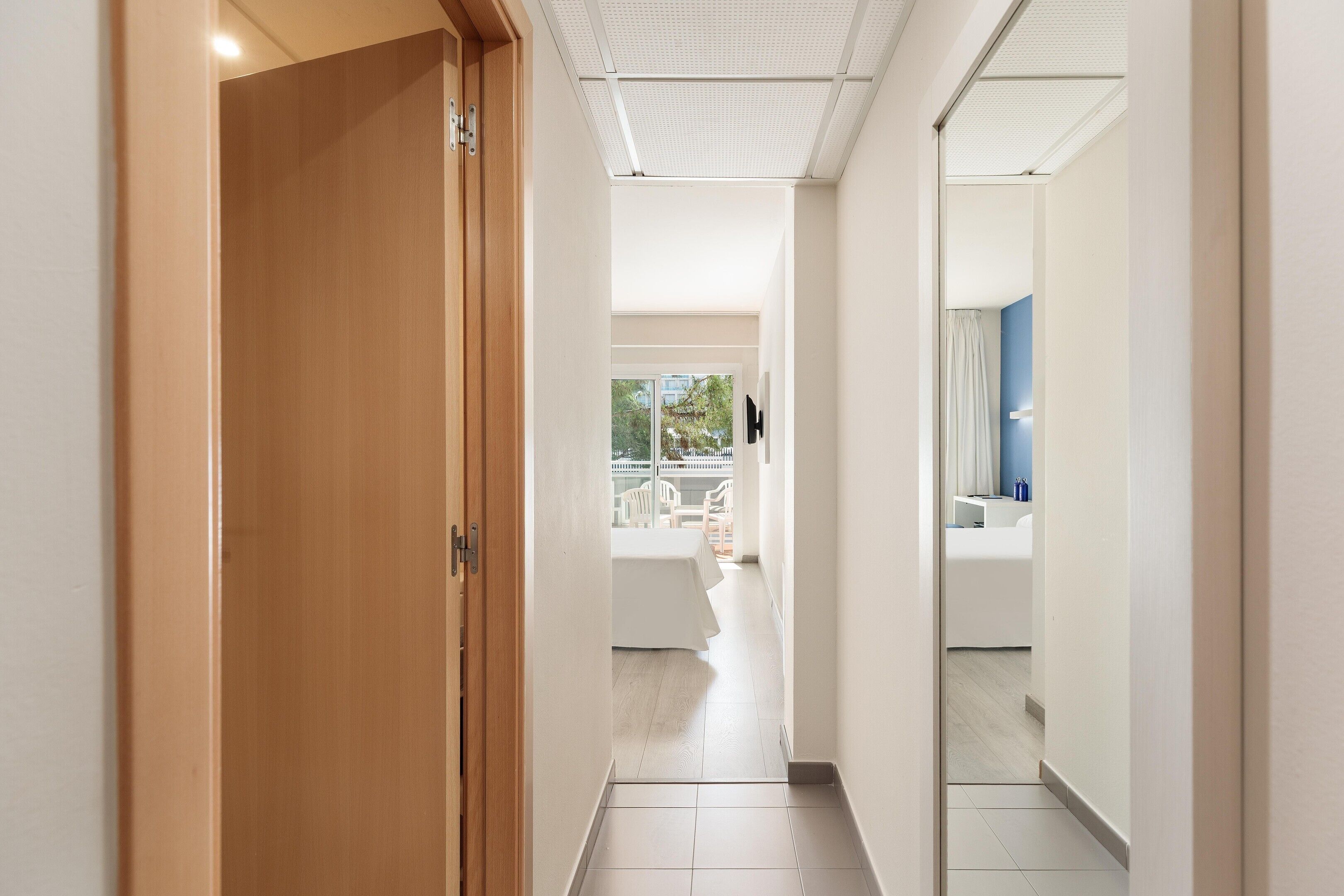 Un pasillo de hotel con una puerta de madera, una pared blanca y vistas a un balcón con mobiliario exterior.
