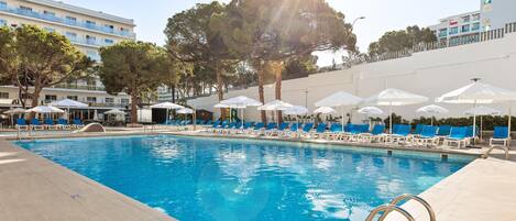 A swimming pool with sunlit water, surrounded by lounge chairs and umbrellas, with buildings in the background.