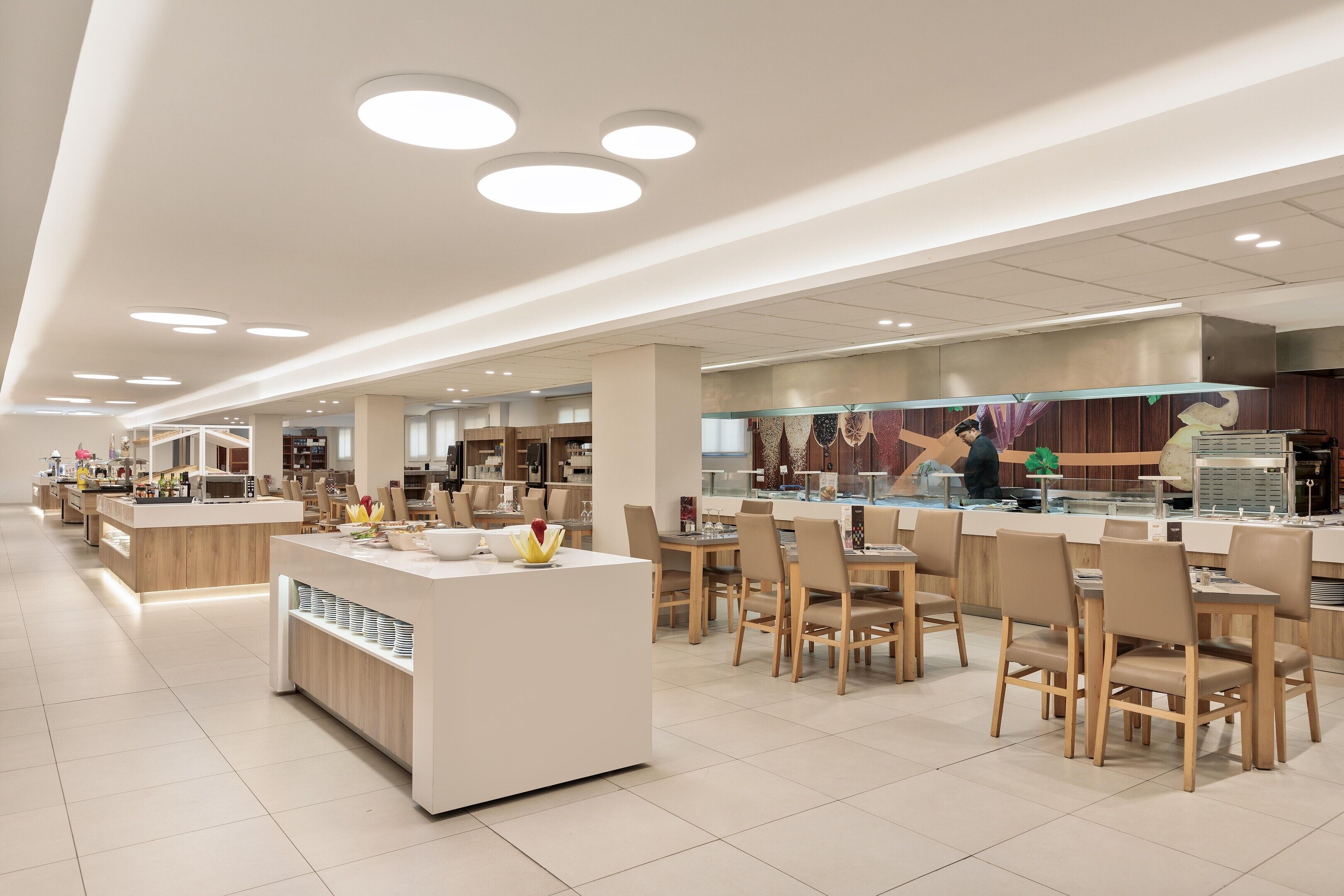 Un café moderno con varias zonas de estar, una barra central con exhibición y un área de cocina al fondo.