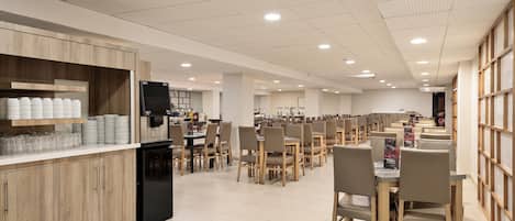 A spacious dining area with multiple tables and chairs, a kitchen area with a refrigerator and shelves stocked with plates and glasses, and a ceiling with recessed lighting.