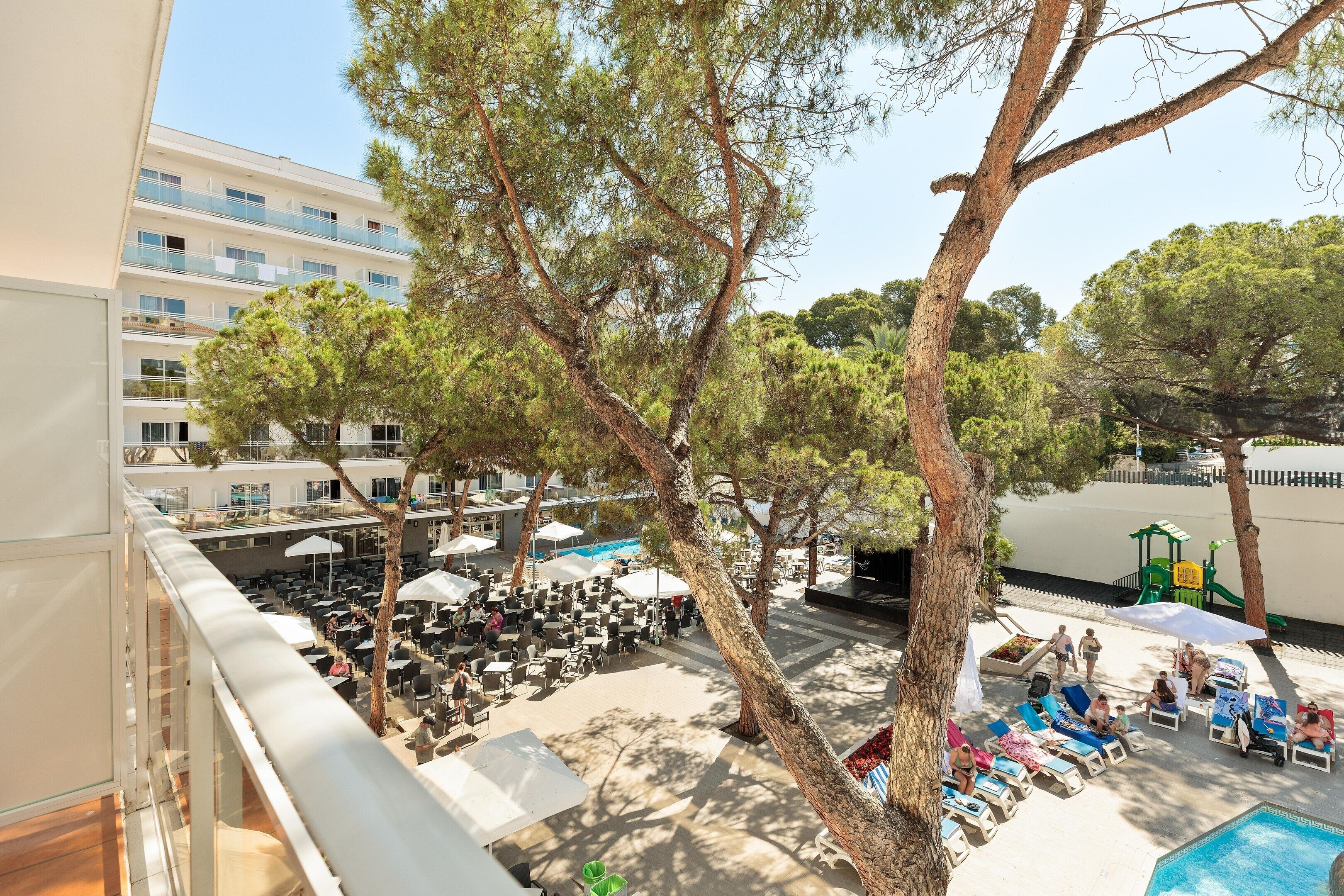 Zona de piscina con sillas de descanso y sombrillas, un edificio con balcones al fondo y árboles que dan sombra.