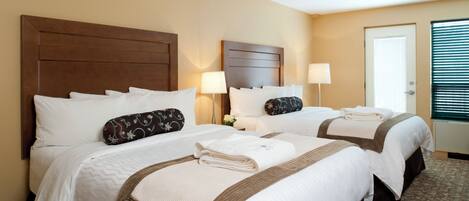 Habitación de hotel con dos camas, cada una con cabecero de madera, ropa de cama blanca y detalles en tono café. Hay una mesita de noche con una lámpara y un jarrón con flores.