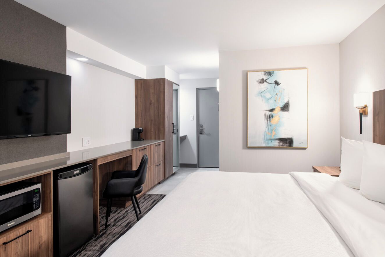Quarto de hotel moderno com cama, escrivaninha com cadeira, micro-ondas, geladeira e televisão de tela plana fixada na parede.