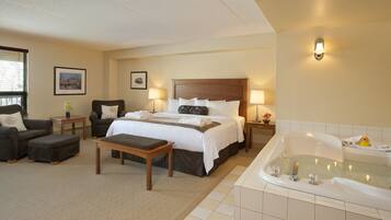 A hotel room with a large bed, a sitting area with a chair and ottoman, a small table, a nightstand with a lamp, a bathroom with a bathtub, and framed pictures on the wall.
