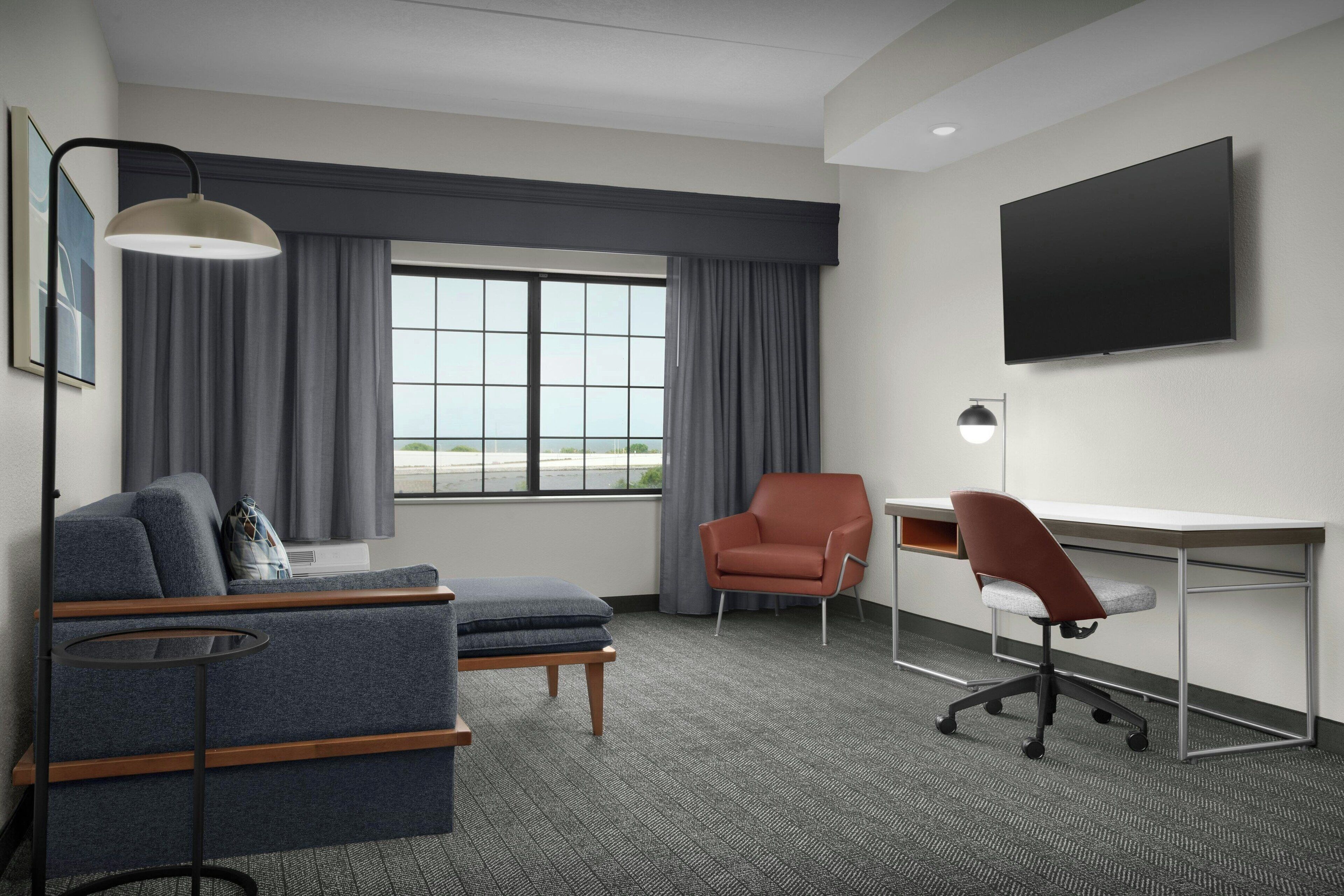 A hotel room with a sofa, a desk, a chair, a TV, and a window with curtains.