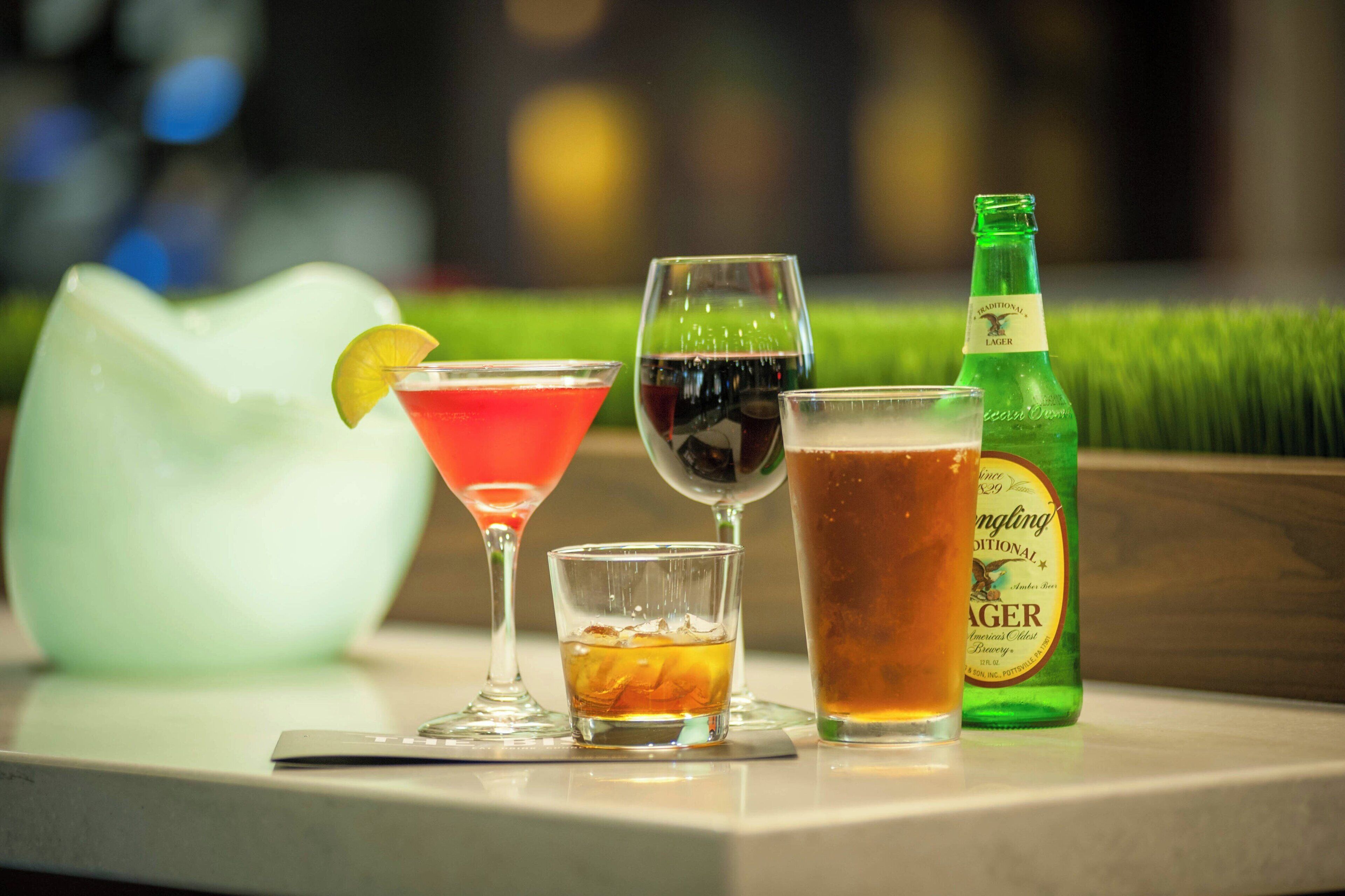 A variety of drinks on a bar counter, including a green bottle of beer, a red cocktail, and a glass of beer with a lime wedge.