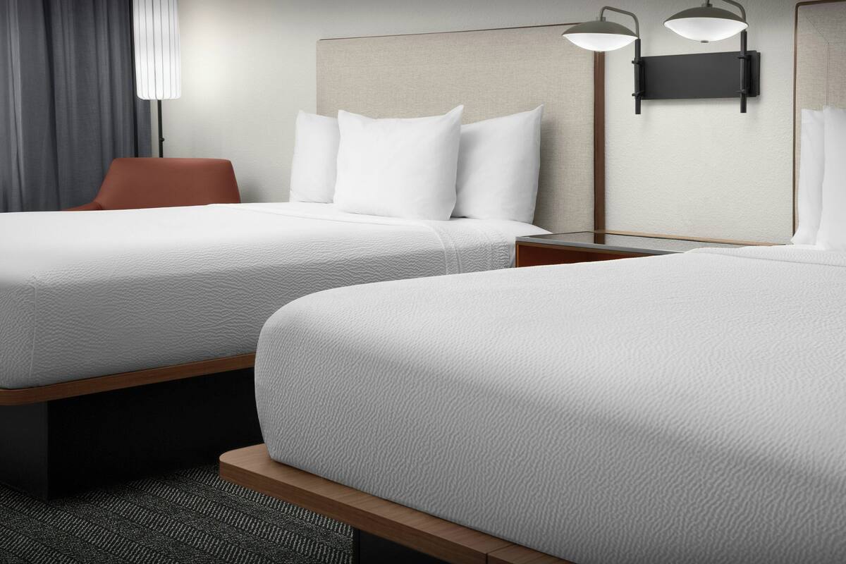 A hotel room with two beds, a nightstand, a lamp, and a chair.