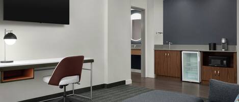 A modern hotel room with a flat-screen TV, a desk with a chair, a kitchenette with a fridge and microwave, and a sofa.