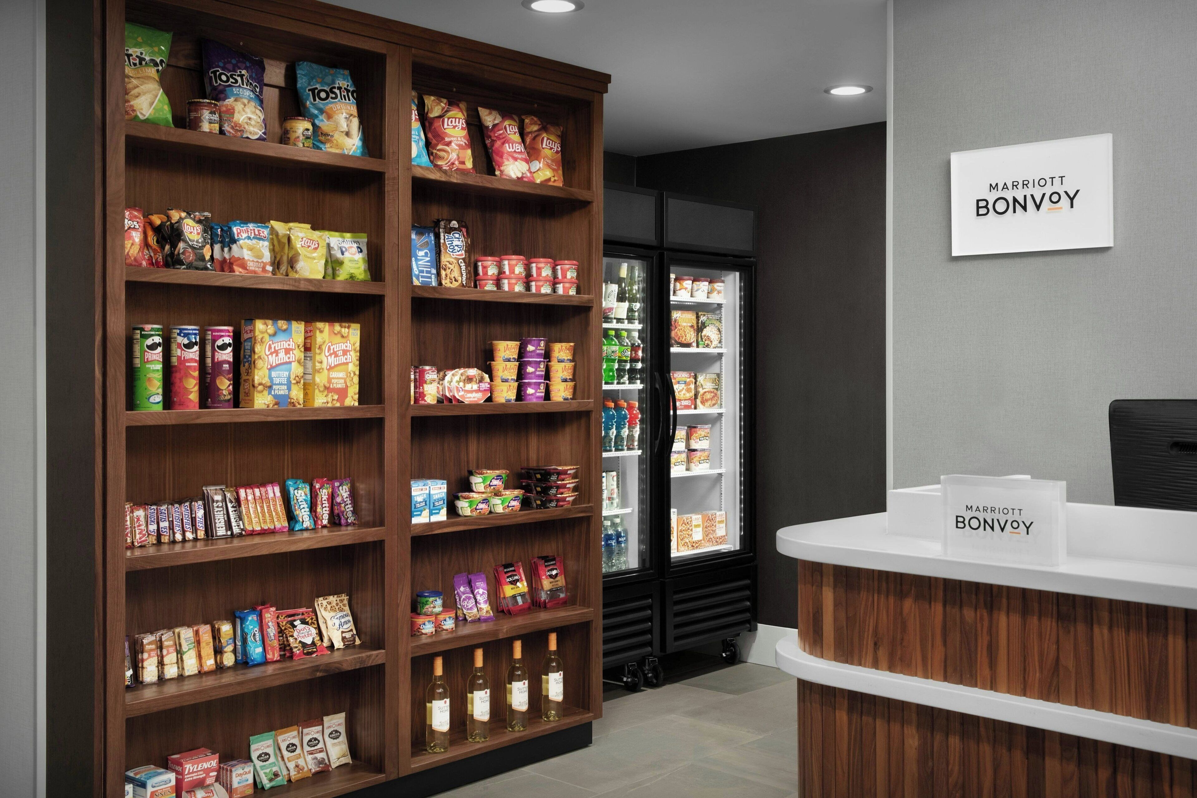 Zona de recepción de hotel con un estante de aperitivos surtido y un refrigerador que exhibe bebidas y alimentos.