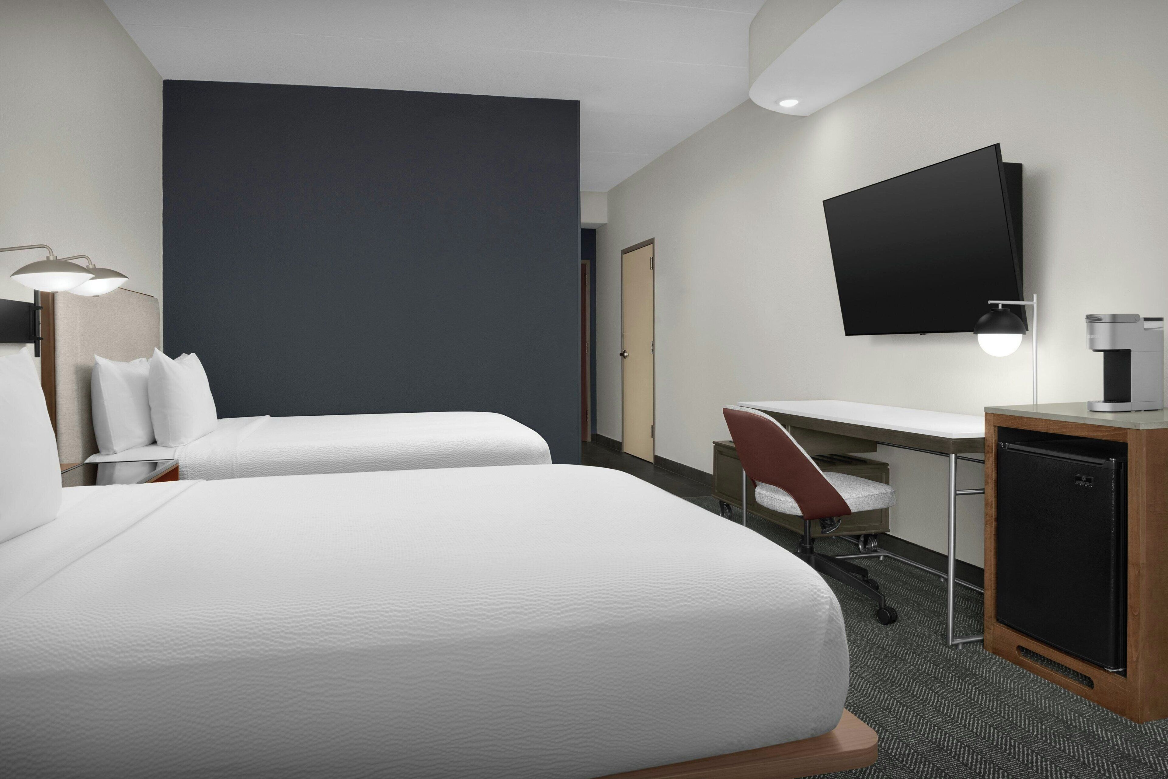 A hotel room with two beds, a desk, a chair, a TV, and a minibar.