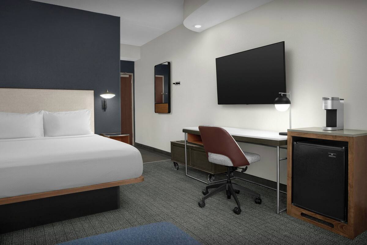 A hotel room with a bed, desk, chair, and a small refrigerator.