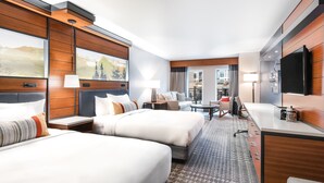 A hotel room with two beds, a desk, a TV, and a view of the city.