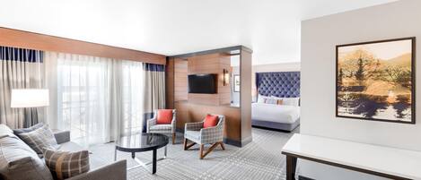 A hotel room with a sofa, two armchairs, a small table, a bed, a TV, and a painting on the wall.