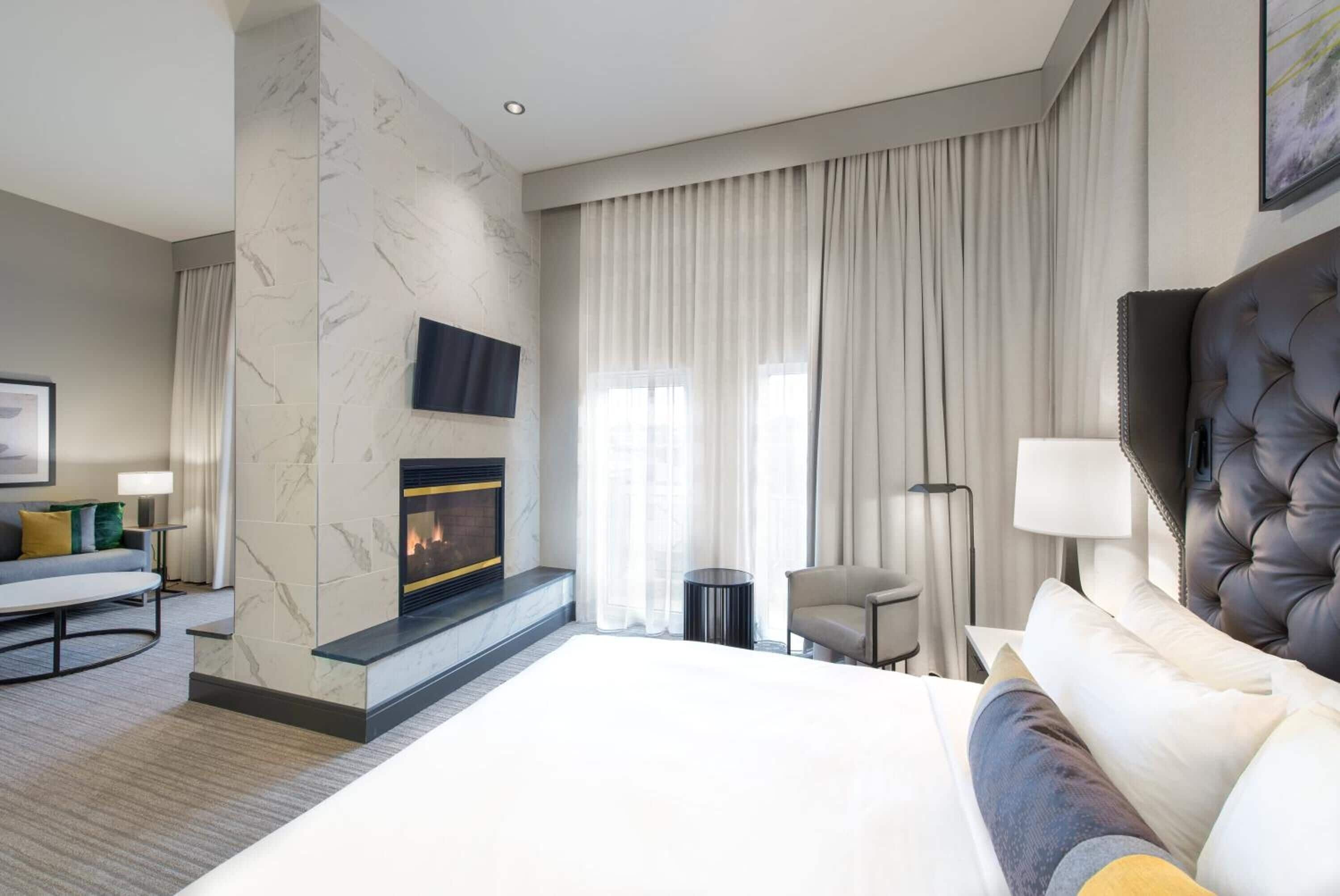 A modern hotel room with a large bed, a fireplace, a television, and a view of the city through large windows.