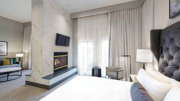 A modern hotel room with a large bed, a fireplace, a television, and a view of the city through large windows.